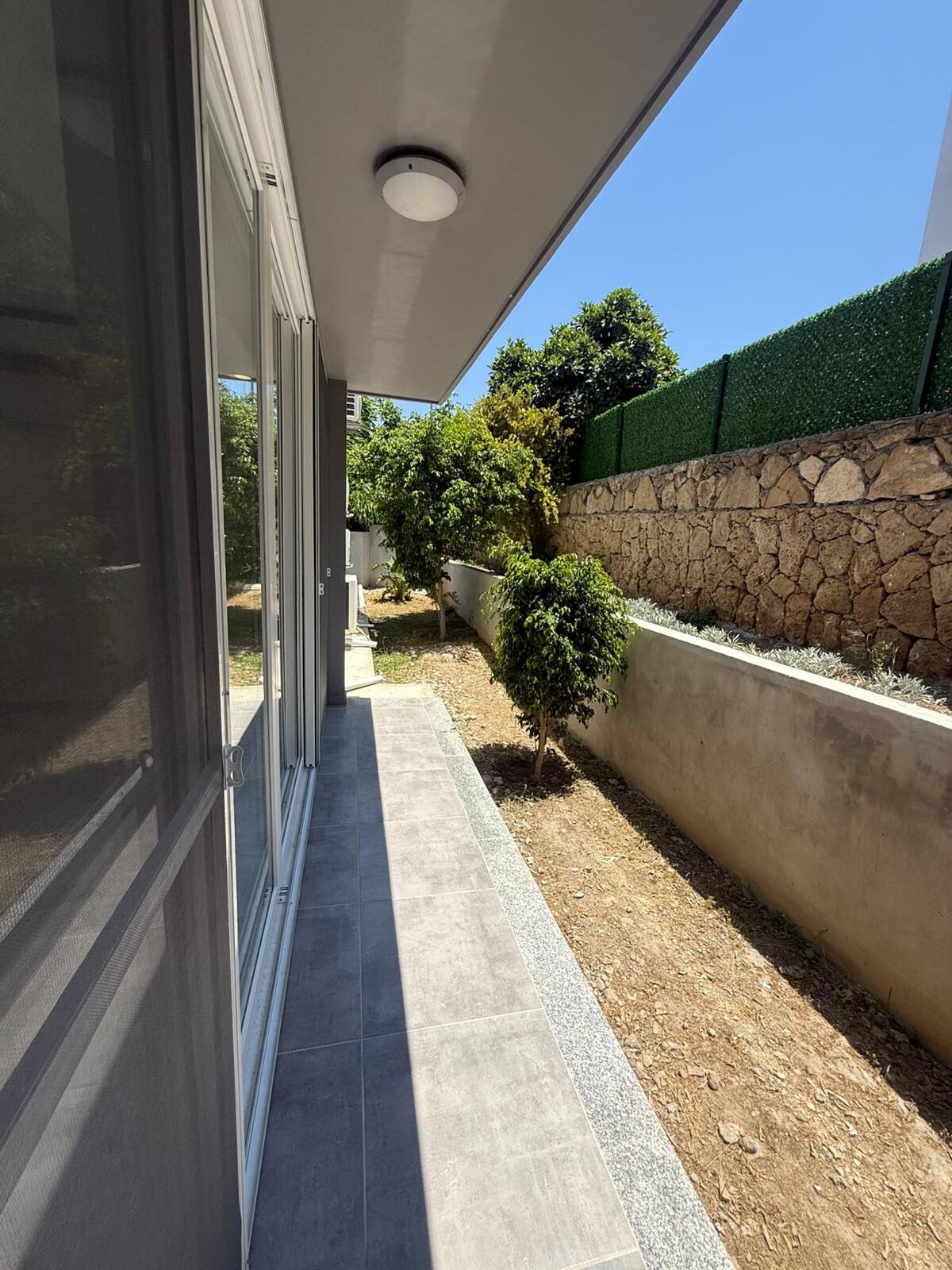 Side terrace along the apartment with young trees and a stone wall